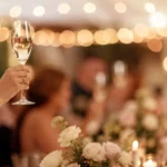 Main levant une flûte de champagne pour porter un toast lors d'une réception de mariage, guirlandes lumineuses en arrière-plan