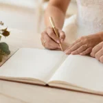 Couple de mariés signant le registre de mariage avec un stylo doré, alliances aux doigts et bouquet de roses posé à côté